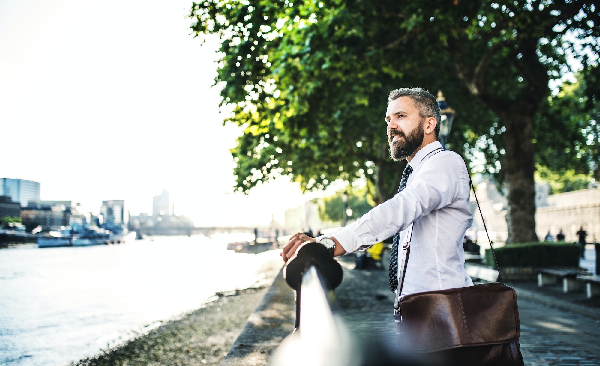 Hipster businessman with laptop bag standing by the river in London.